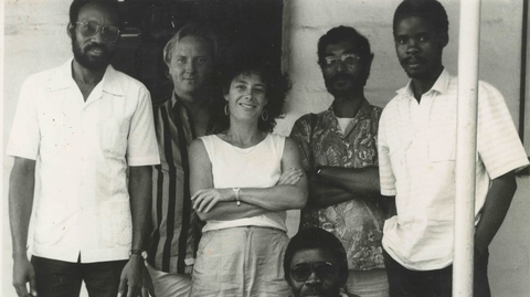 Black-and-white photo of six people, five standing and one sitting, posing casually in front of a plain wall and door.