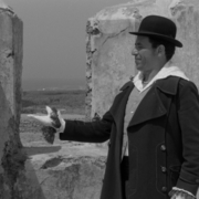 Two men in coats stand near weathered stone walls; one gestures with a gloved hand while the other listens attentively in a black-and-white scene.