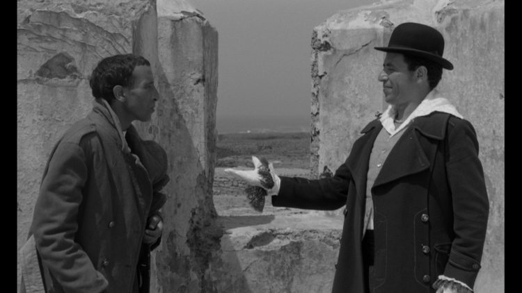 Two men in coats stand near weathered stone walls; one gestures with a gloved hand while the other listens attentively in a black-and-white scene.