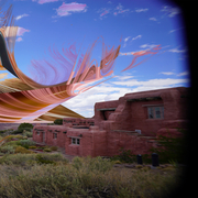 Surreal image with pink-orange abstract shape over a desert landscape, featuring adobe-style buildings and a blue sky with clouds. Dreamlike and vibrant.
