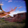 Surreal image with pink-orange abstract shape over a desert landscape, featuring adobe-style buildings and a blue sky with clouds. Dreamlike and vibrant.