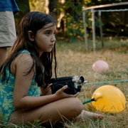 Child in a blue swimsuit sitting on grass holding a vintage video camera, nearby yellow and pink balloons and barefoot people standing.