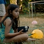 Child in a blue swimsuit sitting on grass holding a vintage video camera, nearby yellow and pink balloons and barefoot people standing.