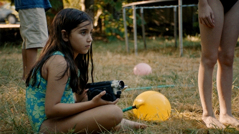 Child in a blue swimsuit sitting on grass holding a vintage video camera, nearby yellow and pink balloons and barefoot people standing.