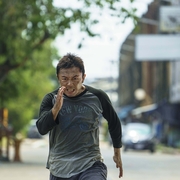 Man in grey and black shirt sprinting on a city street lined with trees and buildings in the background.