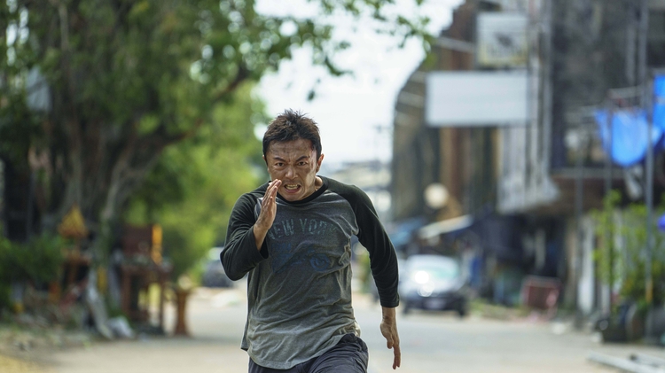 Man in grey and black shirt sprinting on a city street lined with trees and buildings in the background.