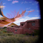Surreal landscape with abstract, swirling shapes in the sky over a desert scene. Red adobe building and vegetation add contrast under a bright blue sky.