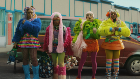Four people wearing colourful, furry jackets, skirts, and boots stand in front of a brown car in an urban parking lot.