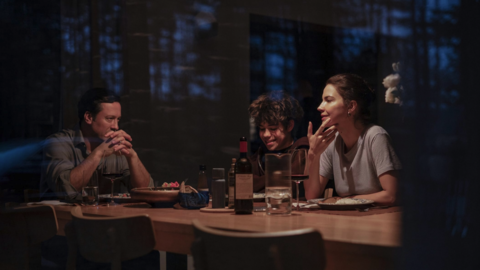 Three people sharing a meal with wine at a wooden table in a dimly lit, cozy indoor setting at night.