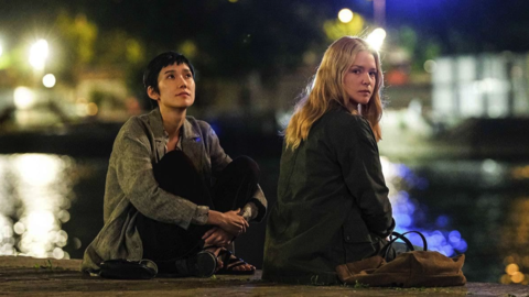 Two people sitting by a waterfront at night, with blurred city lights reflecting on the water in the background.