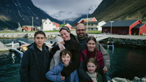 Family group of seven standing by a fjord with boats, red houses, and snow-capped mountains in the background.