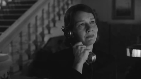 Black and white image of a person in dark clothing holding a vintage telephone handset near their mouth indoors with a staircase in the background.