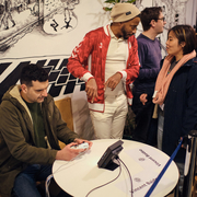 A man plays video games at a round table, while a person in a red jacket and beanie talks to a smiling woman. The background features a detailed mural.