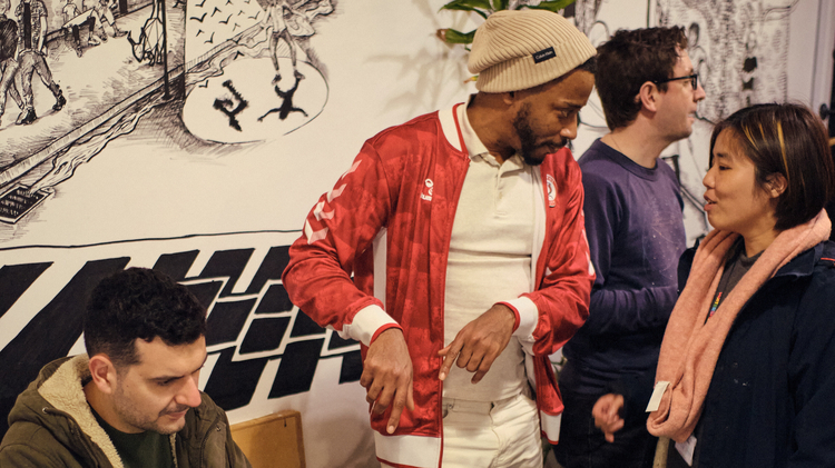A man plays video games at a round table, while a person in a red jacket and beanie talks to a smiling woman. The background features a detailed mural.
