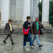 Three people walk on wet stone pavement near large white columns outside a classical-style building.