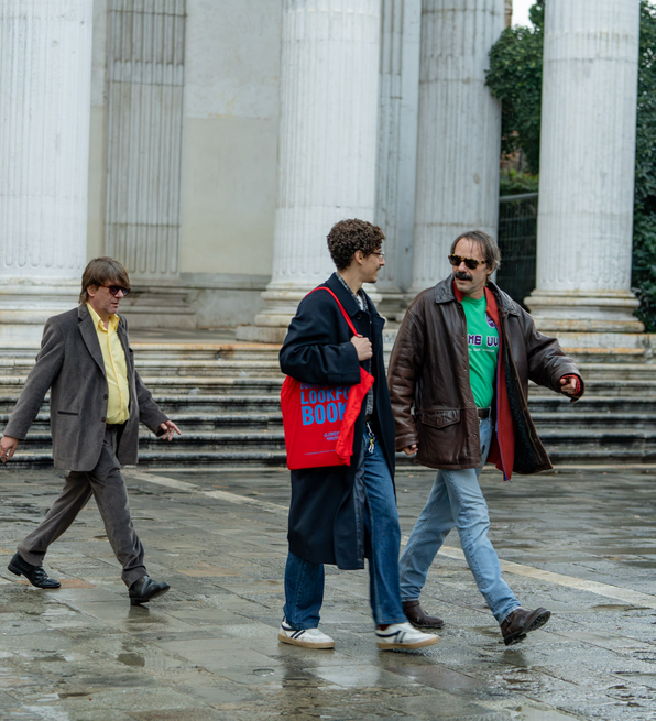 Three people walk on wet stone pavement near large white columns outside a classical-style building.