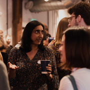 People engaged in conversation at an indoor social gathering, holding drinks and casually interacting in a warmly lit space.