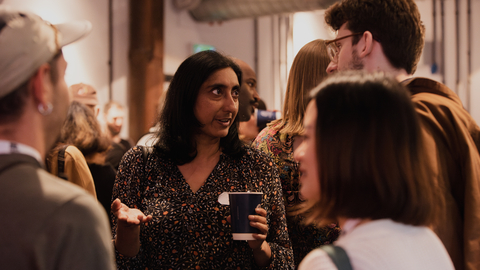 People engaged in conversation at an indoor social gathering, holding drinks and casually interacting in a warmly lit space.