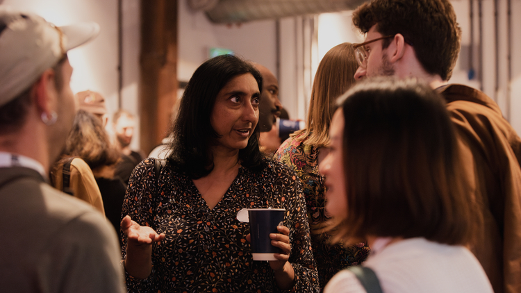People engaged in conversation at an indoor social gathering, holding drinks and casually interacting in a warmly lit space.