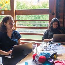 Two brown skinned people work on laptops at a table by a large window with sticky notes. Yarn and crafting supplies are scattered around, creating a creative atmosphere.