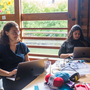 Two brown skinned people work on laptops at a table by a large window with sticky notes. Yarn and crafting supplies are scattered around, creating a creative atmosphere.