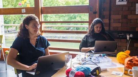 Two brown skinned people work on laptops at a table by a large window with sticky notes. Yarn and crafting supplies are scattered around, creating a creative atmosphere.