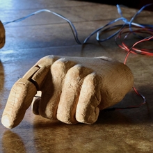 Wooden robotic hands rest on a rustic table. The hands, intricately carved, are connected to wires, suggesting technology and craftsmanship.