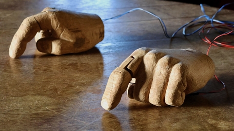 Wooden robotic hands rest on a rustic table. The hands, intricately carved, are connected to wires, suggesting technology and craftsmanship.