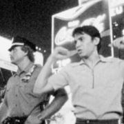 Black and white photo of two men posing confidently in front of illuminated Coca-Cola and other billboards at night.