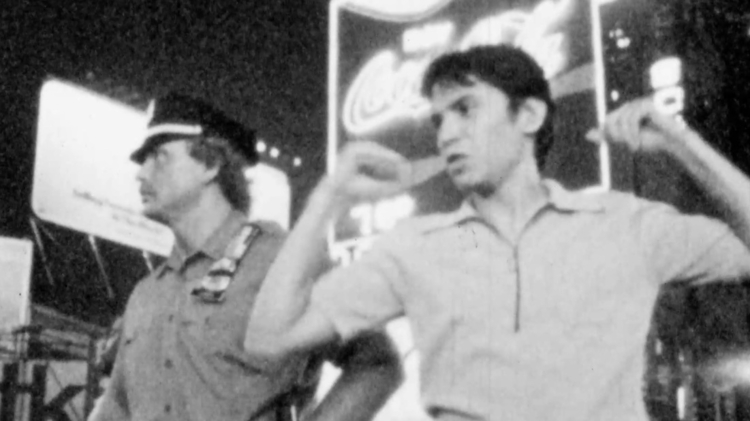 Black and white photo of two men posing confidently in front of illuminated Coca-Cola and other billboards at night.