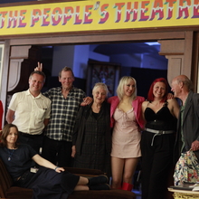 Group of people posing under a colourful "The People's Theatre" sign, with one person reclining on a vintage sofa outside a theatre lobby.