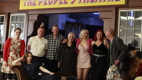 Group of people posing under a colourful "The People's Theatre" sign, with one person reclining on a vintage sofa outside a theatre lobby.