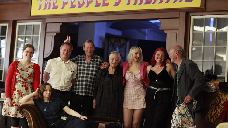 Group of people posing under a colourful "The People's Theatre" sign, with one person reclining on a vintage sofa outside a theatre lobby.