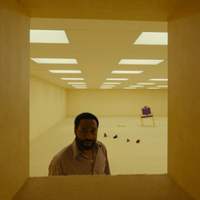 Man stands in beige room with fluorescent ceiling lights, looking through a small rectangular opening; scattered shoes and a purple chair in background.
