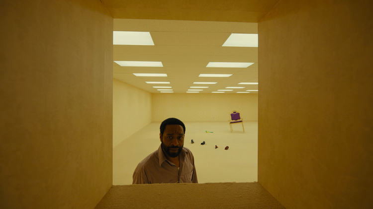 Man stands in beige room with fluorescent ceiling lights, looking through a small rectangular opening; scattered shoes and a purple chair in background.
