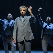 Male performer in grey suit holds up four fingers, flanked by two background dancers in suits striking expressive poses on stage.