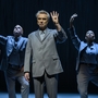 Male performer in grey suit holds up four fingers, flanked by two background dancers in suits striking expressive poses on stage.