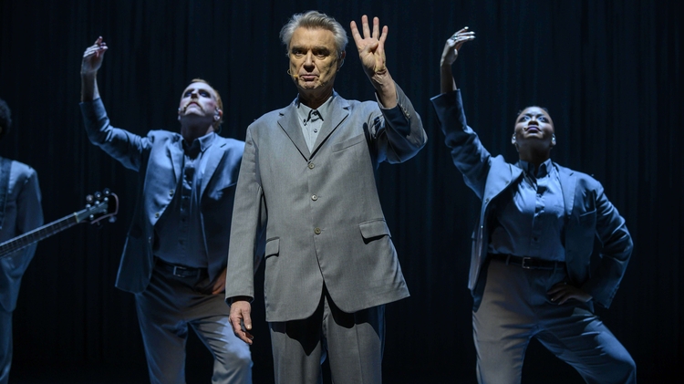 Male performer in grey suit holds up four fingers, flanked by two background dancers in suits striking expressive poses on stage.