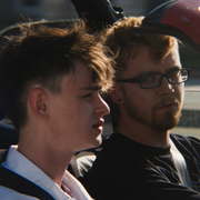 Two young men sitting side by side in a car, both wearing seat belts, with the driver wearing glasses and looking forward.