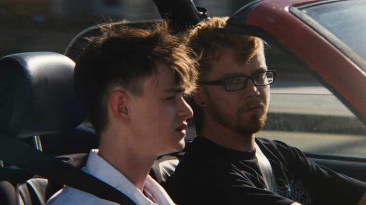 Two young men sitting side by side in a car, both wearing seat belts, with the driver wearing glasses and looking forward.