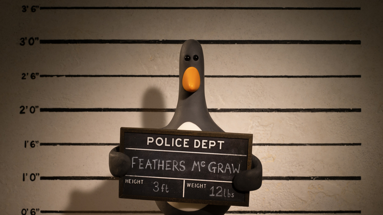 Penguin holding a police mugshot board in front of a height chart, named Feathers McGraw, 3 ft tall and weighing 12 lbs.