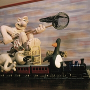 Claymation characters, including a man with a net, a dog, and a penguin, riding a toy train on tracks indoors.
