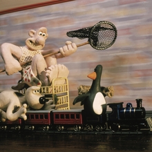 Claymation characters, including a man with a net, a dog, and a penguin, riding a toy train on tracks indoors.