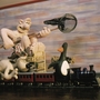 Claymation characters, including a man with a net, a dog, and a penguin, riding a toy train on tracks indoors.