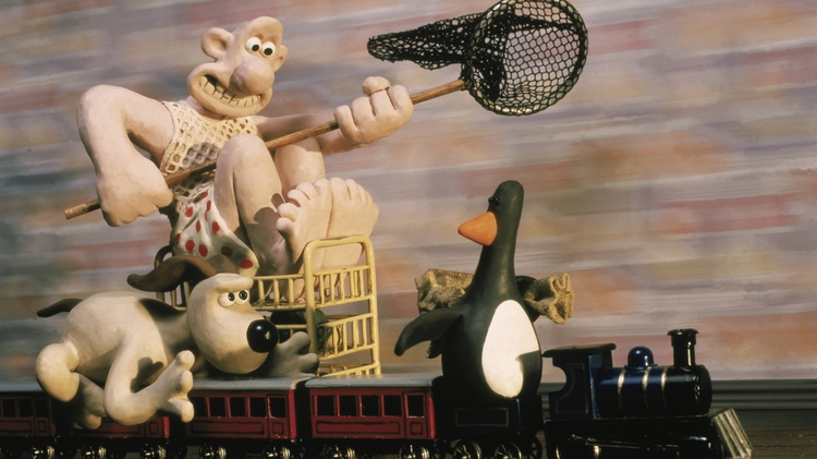 Claymation characters, including a man with a net, a dog, and a penguin, riding a toy train on tracks indoors.