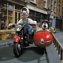 Claymation characters Wallace and Gromit riding a motorcycle with a sidecar on a quiet street with brick buildings.