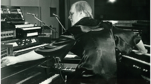 Black and white photo of a man playing multiple keyboards and synthesizers in a studio setting.