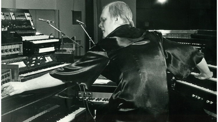 Black and white photo of a man playing multiple keyboards and synthesizers in a studio setting.
