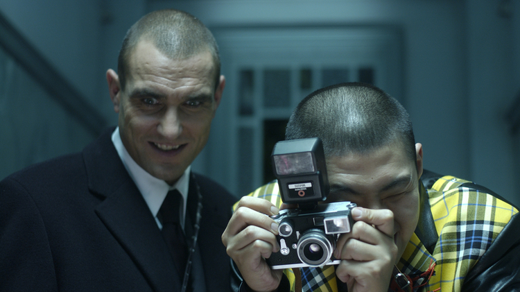 Two men standing next to each other - one taking a photo with a camera