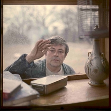 Man with a curious expression touches a dusty window from outside, viewed from inside a room with books and a decorative lamp.